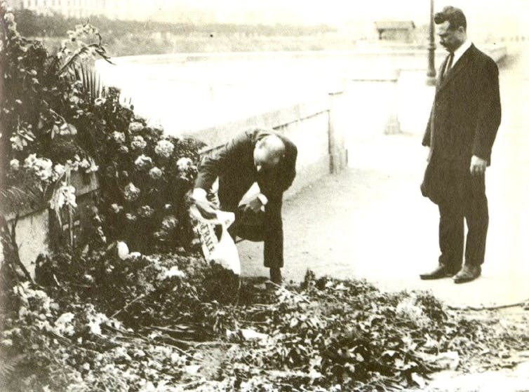 A black and white photo of a man bending down to take a close look at a huge pile of flowers placed in memorial.