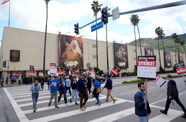 Un grupo de personas portando pancartas caminan por un paso de peatones frente a un gran edificio cubierto con grandes pancartas que contienen imágenes de personas