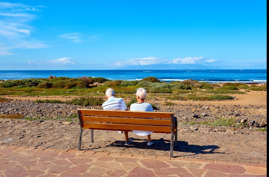 An elderly couple sit on a wooden bench looking out to sea on a bright day. 