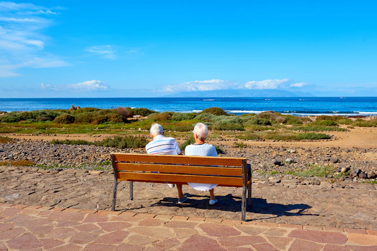 An elderly couple sit on a wooden bench looking out to sea on a bright day.