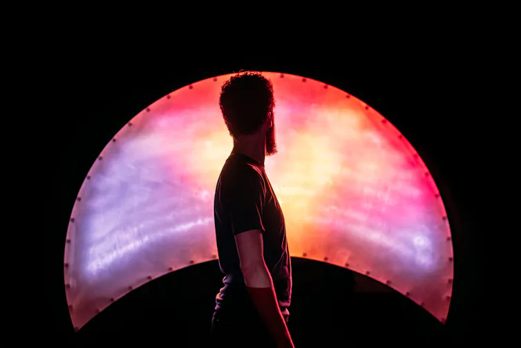 A man on stage facing a light shaped like a crescent moon on its side.