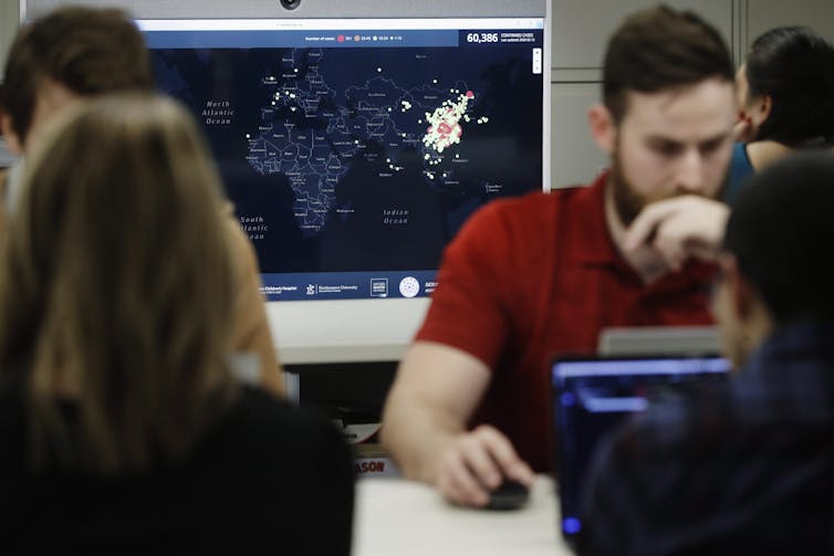 a man sitting at a computer is concentrating on the screen; another screen behind shows a map with a disease outbreak cluster