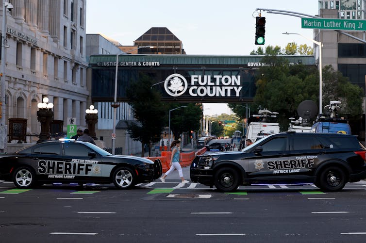Two black cars that say 'sheriff' on it block off a street in front of a walk over that says Fulton County and nearby government buildings.