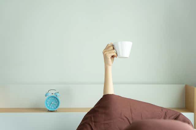 Hand holding coffee cup above comforter, a blue alarm clock resting on the mantle