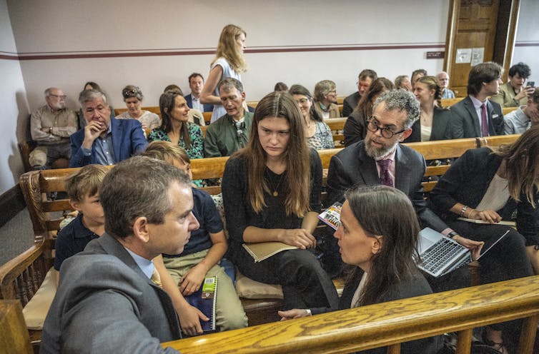 Una joven y dos niños escuchan a los abogados. Los jóvenes llenan dos filas de bancos detrás de ellos en la pequeña sala del tribunal.