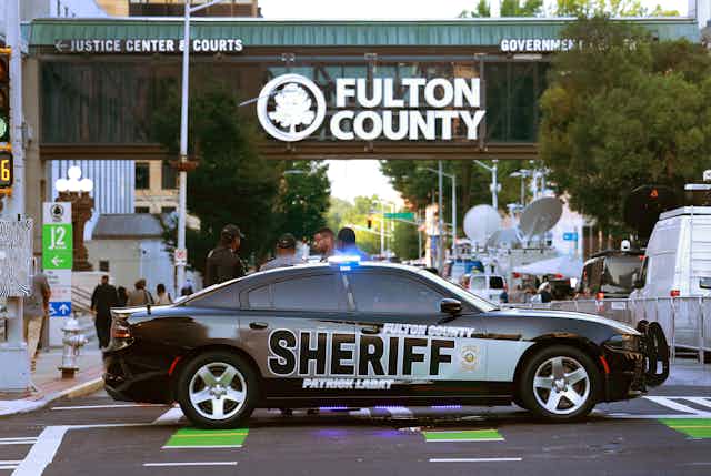 A sheriff's car sits across an intersection in front of an official building with the sign "Fulton County" on it.