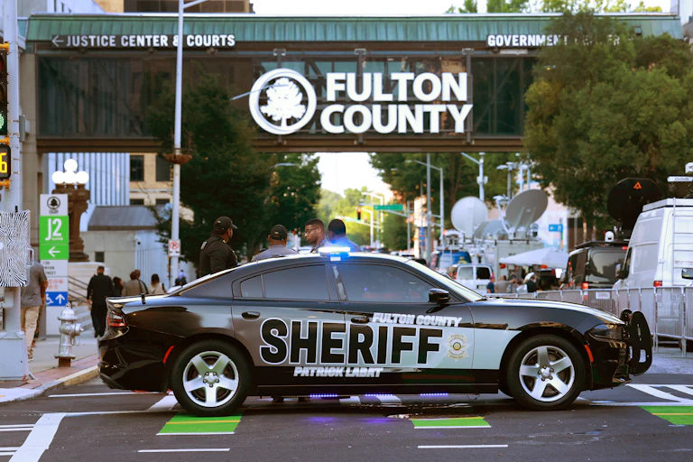 A sheriff's car sits across an intersection in front of an official building with the sign "Fulton County" on it.