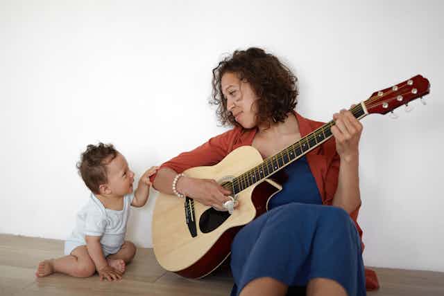 Happy Hispanic woman sitting on floor, playing guitar to her baby.