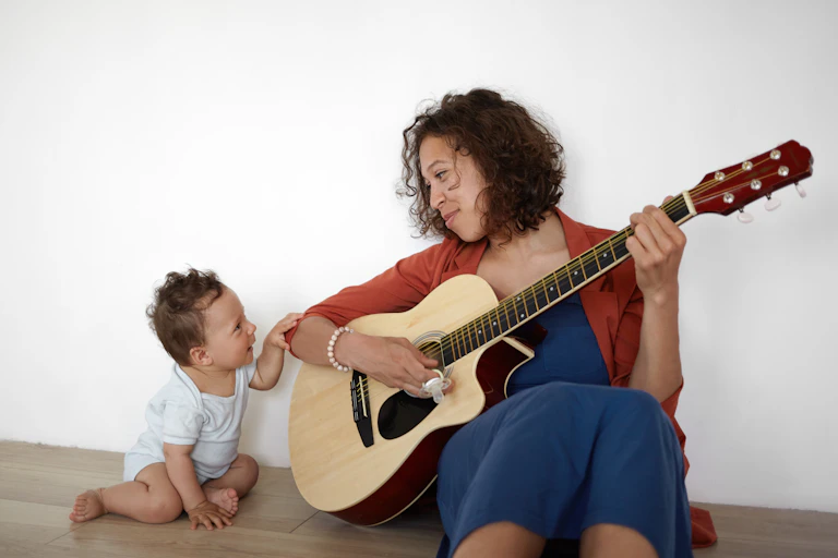 Happy Hispanic woman sitting on floor, playing guitar to her baby.