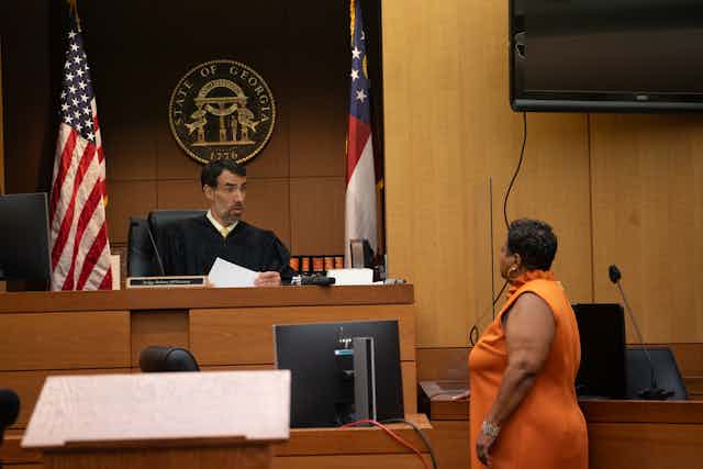 A judge in a black robe appears to review papers and talk with a woman in an orange dress in a U.S. courtroom. 