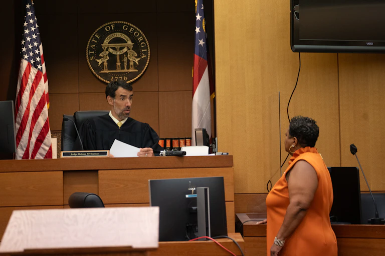 A judge in a black robe appears to review papers and talk with a woman in an orange dress in a U.S. courtroom.