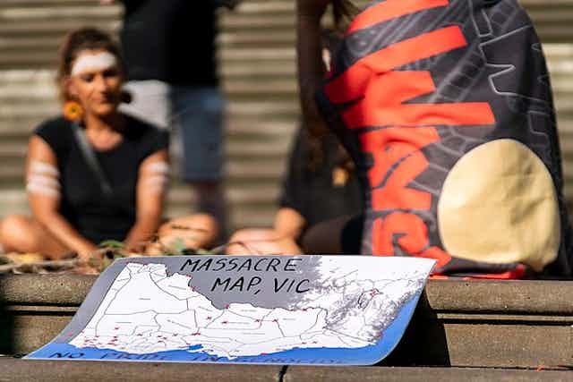 A group of people sit together around a pile of gum leaves, in reflection. They wear ochre across their foreheads. A map is in the foreground that reads 'Massacre Map, VIC'