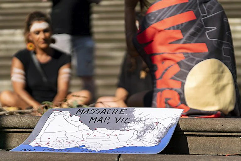A group of people sit together around a pile of gum leaves, in reflection. They wear ochre across their foreheads. A map is in the foreground that reads 'Massacre Map, VIC'