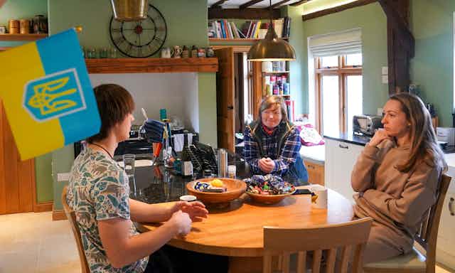 Two Ukrainian refugees, a young man and a middle-aged woman, sit at a table in a cottage-style British kitchen with their host, a middle-aged British woman. A Ukrainian flag is hanging on the wall