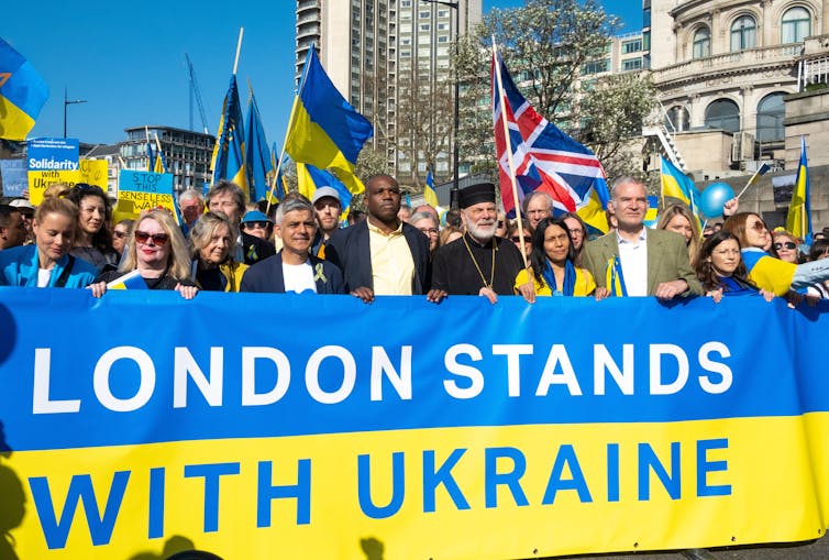 A crowd of people, led by London Mayor Sadiq Khan and others, marches behind a large blue and yellow banner reading 'London Stands With Ukraine'.