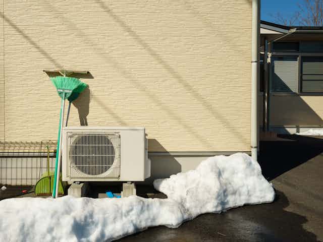 A heat pump attached to the side of a house and surrounded by snow and with a brush propped up next to it.