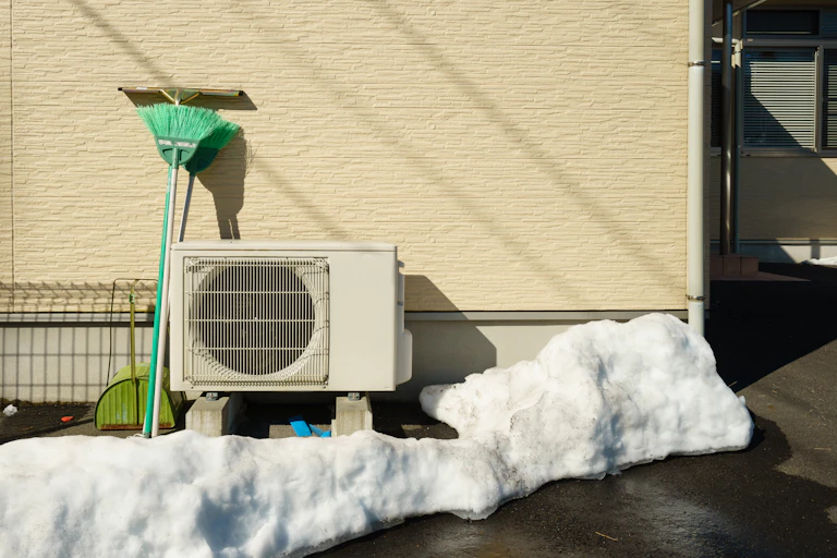 A heat pump attached to the side of a house and surrounded by snow and with a brush propped up next to it.
