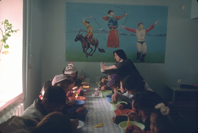 A Catholic nun handing out food to children seated on a rug in two rows.