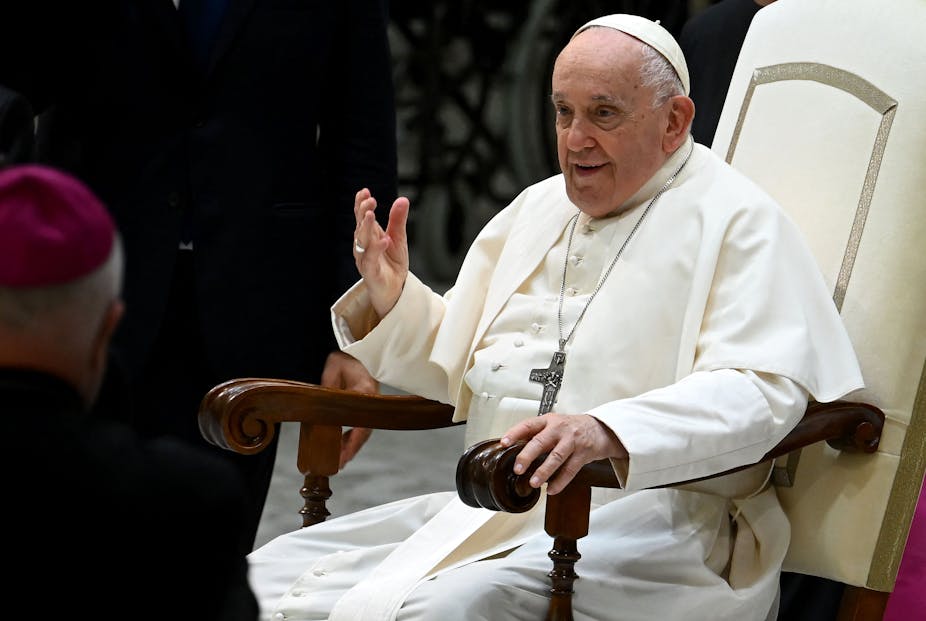 Pope Francis, dressed in a white robe with a crucifix around his neck, gesturing with his hand while talking.