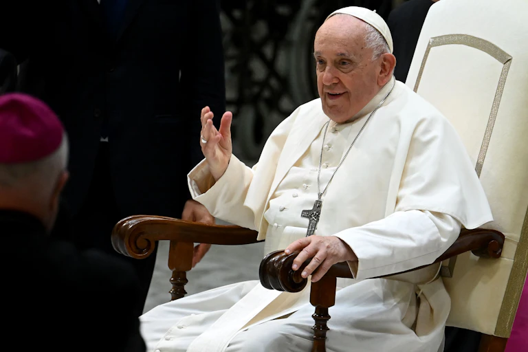 Pope Francis, dressed in a white robe with a crucifix around his neck, gesturing with his hand while talking.