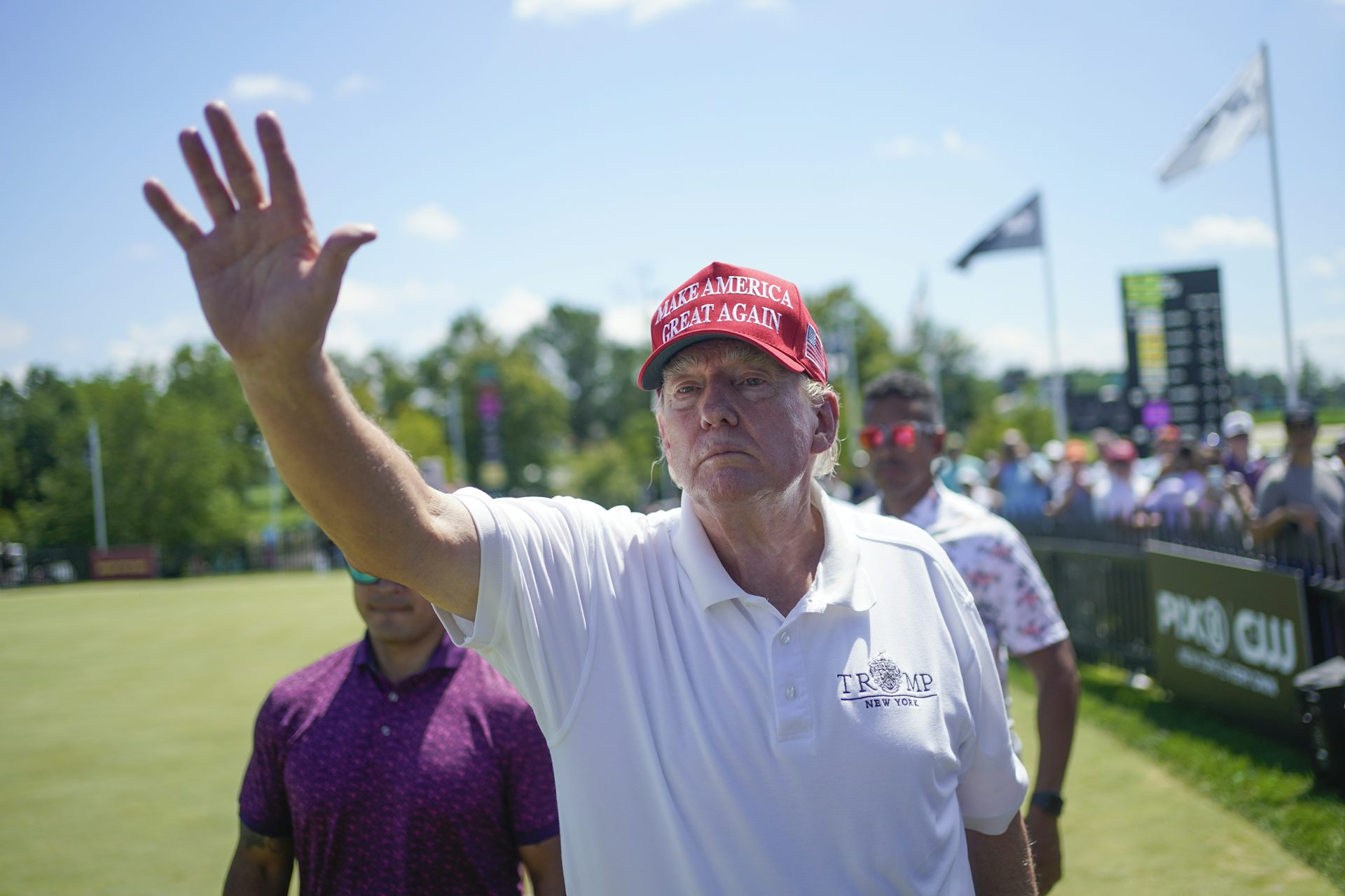 Un homme vêtu d'une chemise de golf blanche et portant une casquette rouge salue sans sourire.