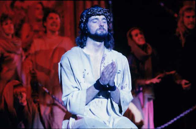 An actor playing Jesus, wearing a white robe and crown of thorns, sits with hands folded in prayer.