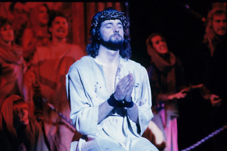 An actor playing Jesus, wearing a white robe and crown of thorns, sits with hands folded in prayer.