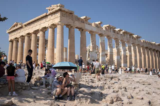 A tourist drinks water as she and a man sit under an umbrella in front of the Parthenon temple.