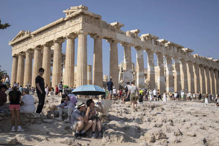 A tourist drinks water as she and a man sit under an umbrella in front of the Parthenon temple.