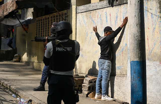 A man stands against the wall with his hands up while armed police stand guard.