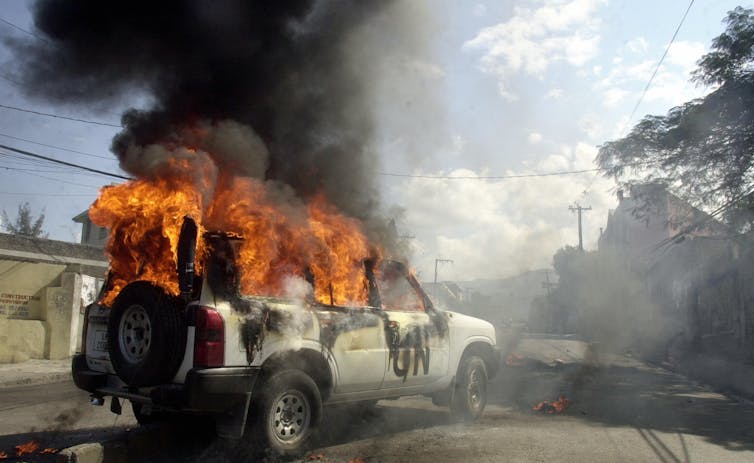 A van with the word “UN” written on the side is on fire and dark smoke is rising from it.