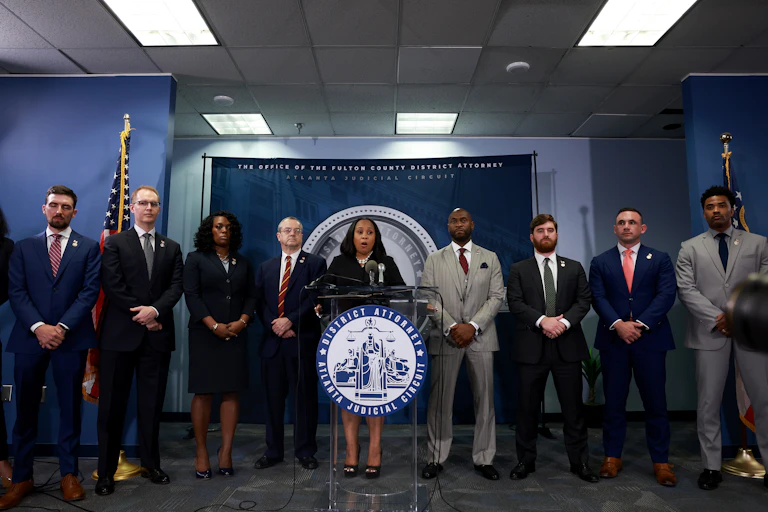 Fani Willis wears a dark outfit and stands in the middle of a row of people in formal clothing at a lectern.