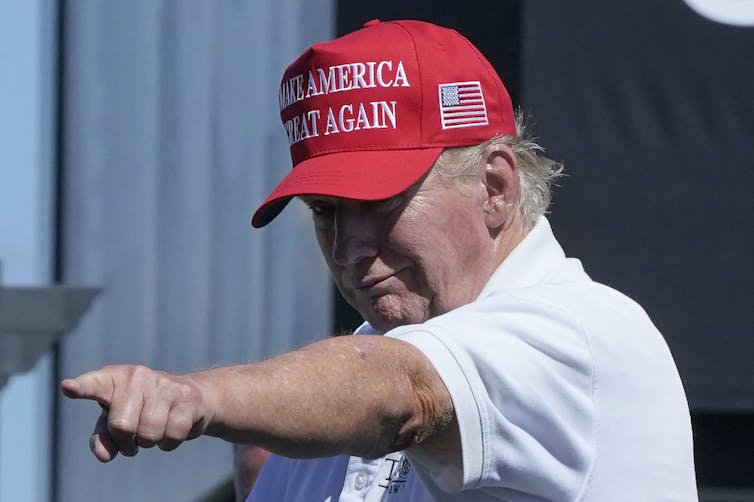 Donald Trump wears a Make America Great Again red hat and a white shirt and points towards the camera.