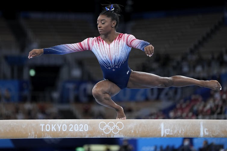A young Black women in a leotard balances on one bent leg while on a balance beam