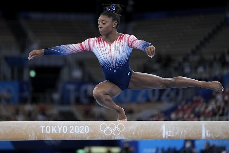 Una joven negra con un maillot hace equilibrios sobre una pierna doblada mientras está en una viga de equilibrio