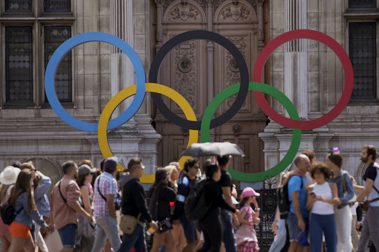 A group of people walk in front of a large Olympic logo sitting in front of a pair of ornate doors