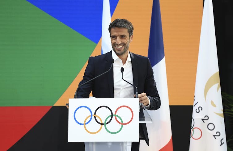 A man in an open-collar shirt and blazer smiles from behind a podium emblazoned with the Olympic logo