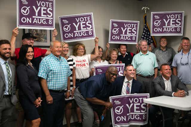 About a dozen people crowded together holding signs to 'Vote YES on Aug. 8'
