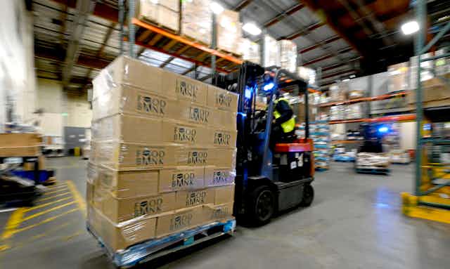 A worker in a safety vest operates heavy equipment to move a large number of boxes around a warehouse.