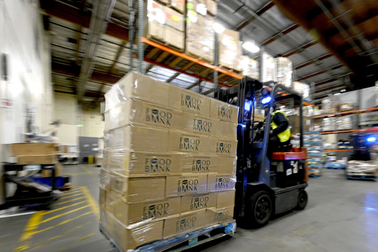 A worker in a safety vest operates heavy equipment to move a large number of boxes around a warehouse.