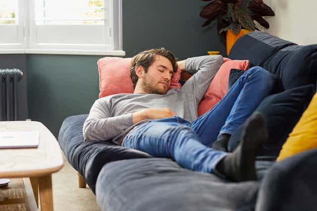 With sunlight streaming through a nearby window, a young man dressed in grey pullover shirt and jeans takes a nap on the couch.