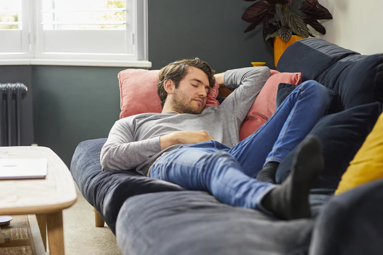 With sunlight streaming through a nearby window, a young man dressed in grey pullover shirt and jeans takes a nap on the couch.