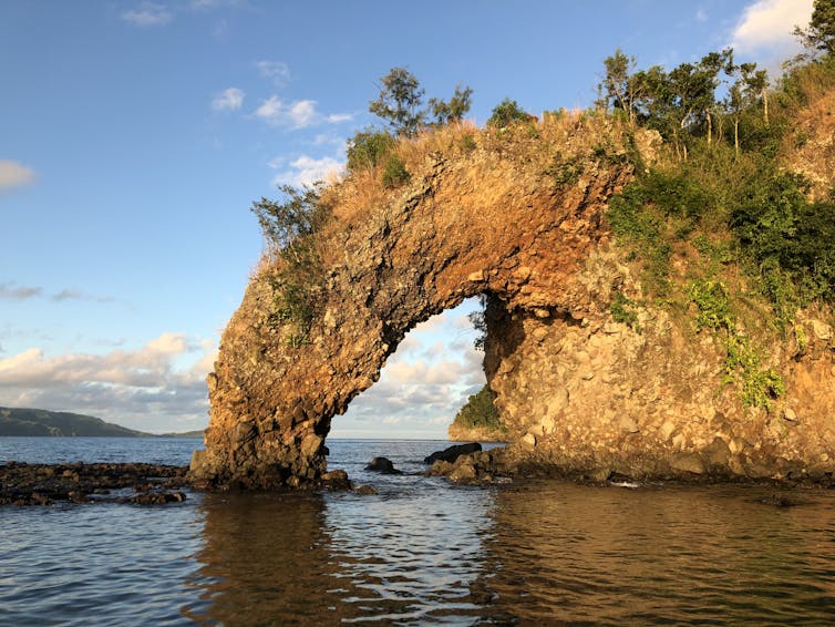 An orange rock jutting out of the water with a large hole within