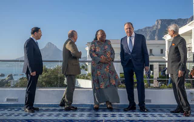 Four men and a woman stand and laugh in front of a white building and mountains.