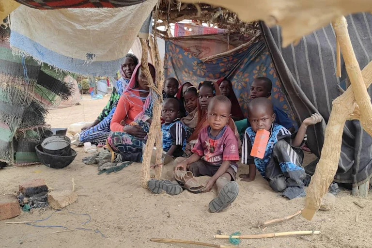 Groupe de gens avec de nombreux jeunes enfants assis par terre sous une tente de fortune