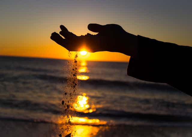 sable glissant des doigts d'une main à contrejour sur un soleil couchant