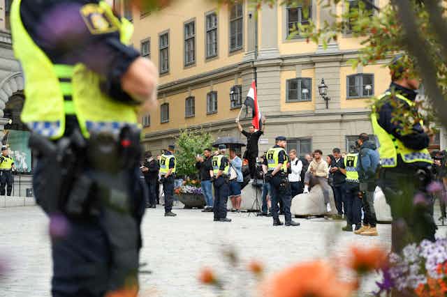 Police officers standing in a circle on a Swedish street next to a man holding up a copy of the Qur'an and what appears to be an Iraqi flag.  