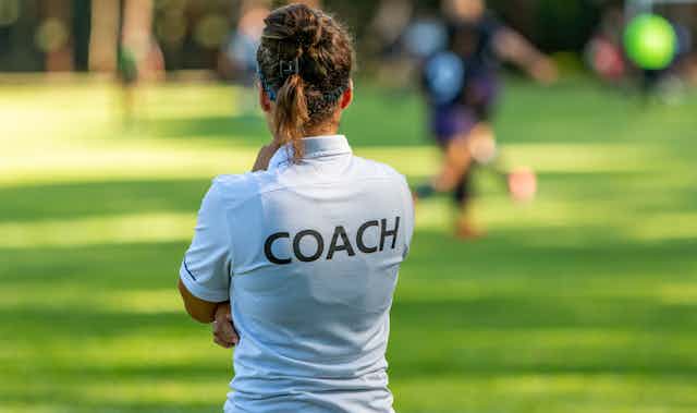 A women wearing a white t-shirt with the word 'Coach' on the back faces a soccer game