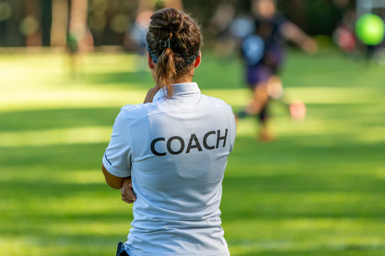 A women wearing a white t-shirt with the word 'Coach' on the back faces a soccer game