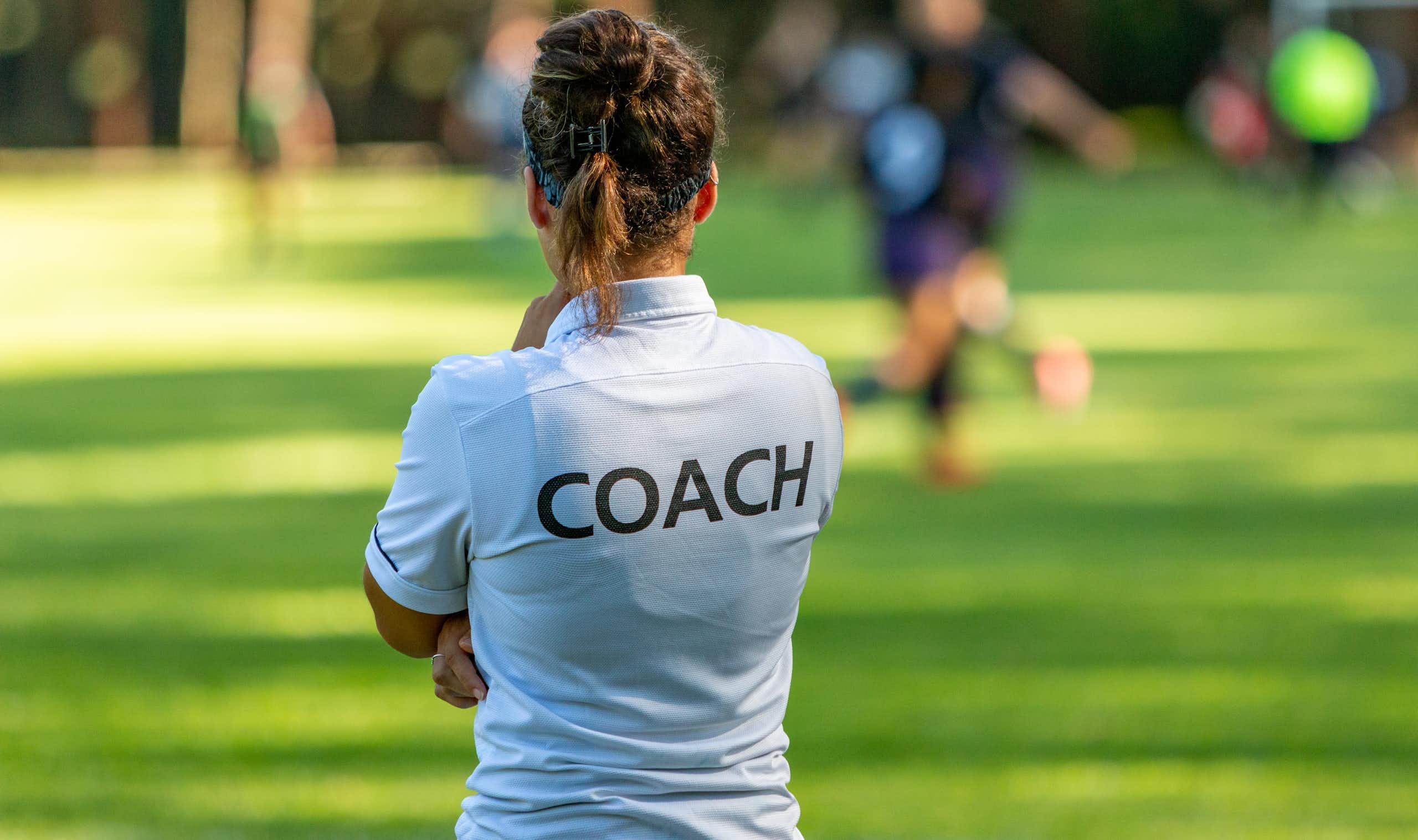 A women wearing a white t-shirt with the word 'Coach' on the back faces a soccer game